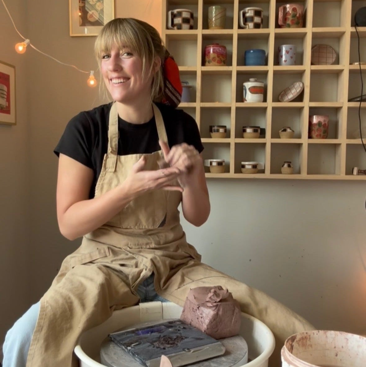Person in a pottery studio with shelves of ceramic items and a potter's wheel.