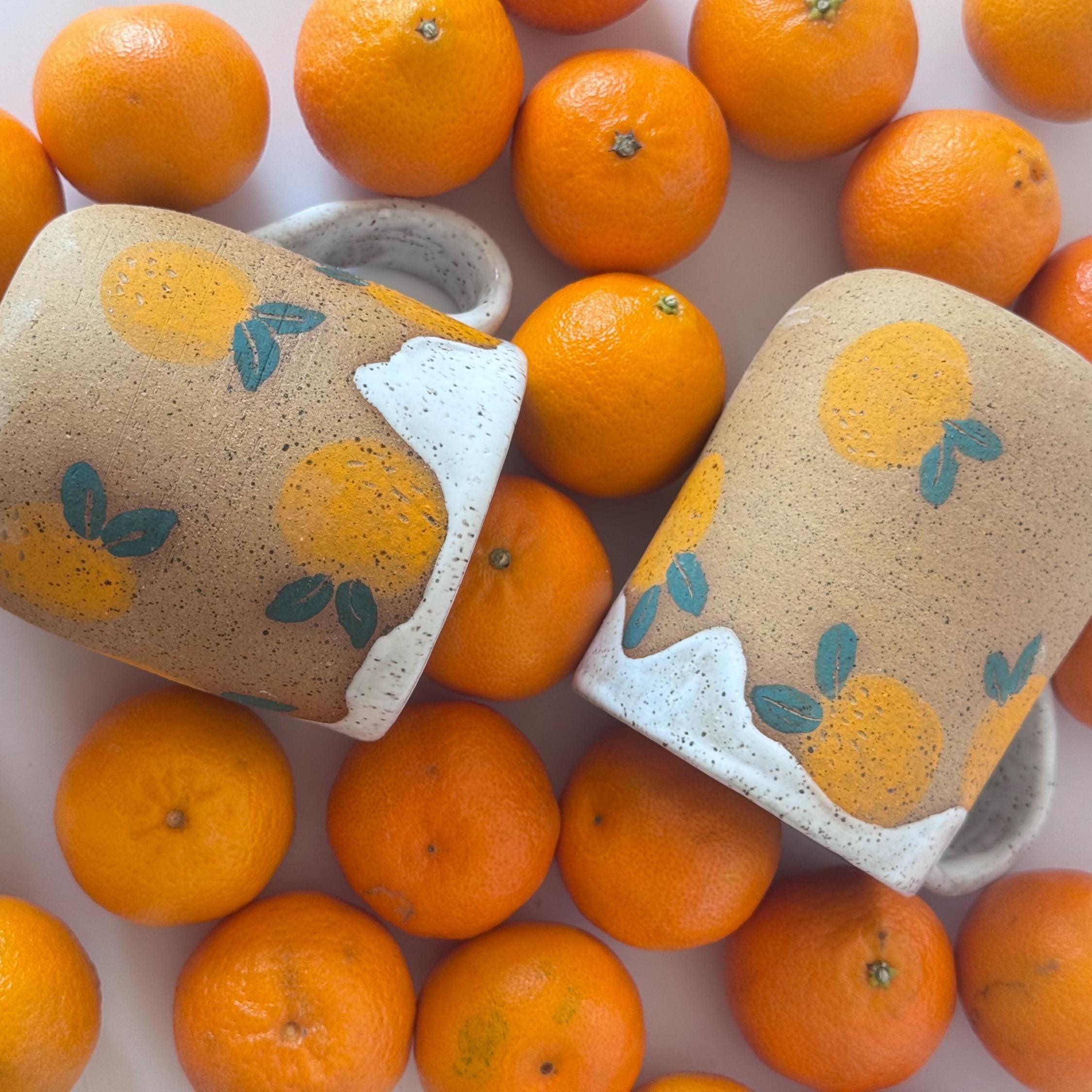 Two ceramic containers with orange designs on a bed of oranges.