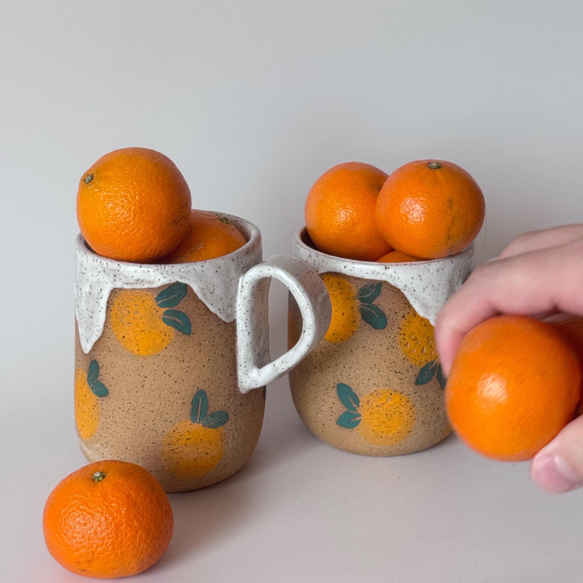 Two ceramic cups with orange designs filled with oranges on a light gray background.