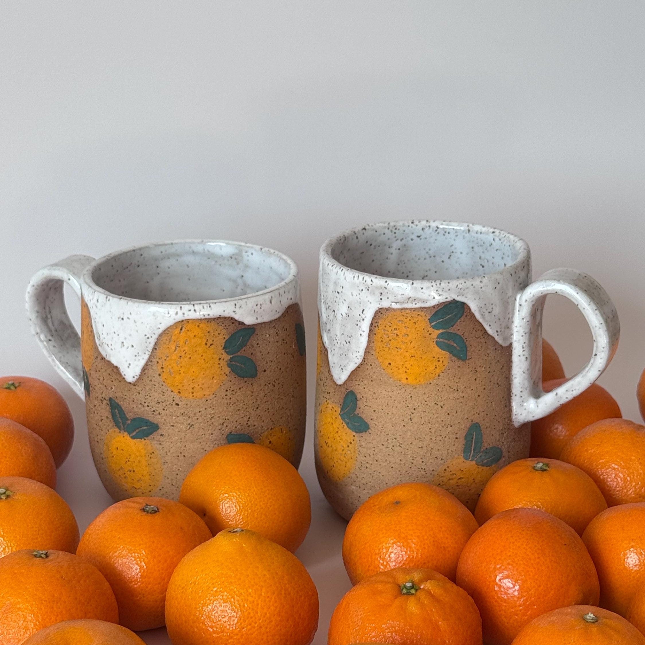 Two ceramic mugs with orange designs surrounded by oranges on a light gray background