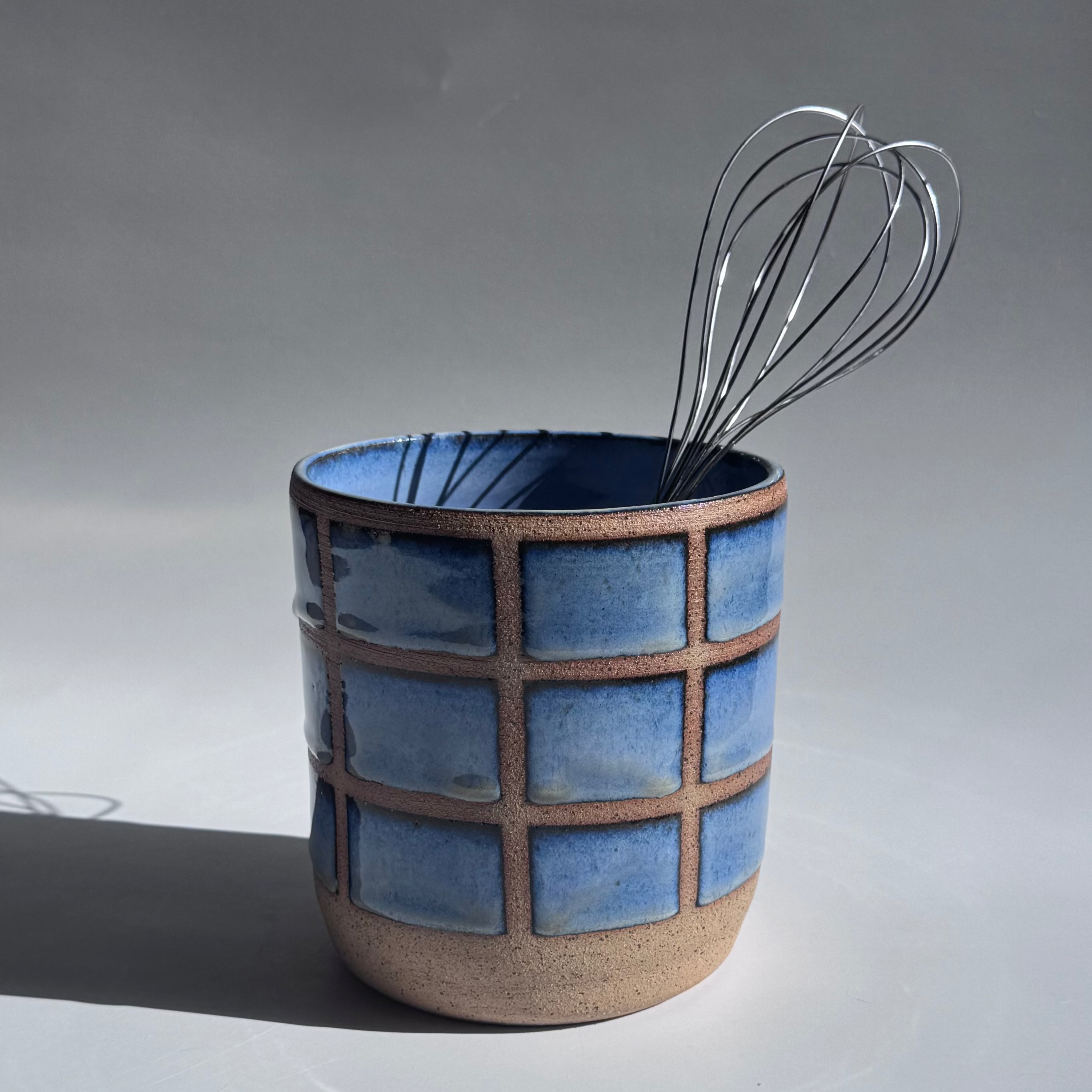 Ceramic vase with blue grid pattern holding a whisk on a gray background