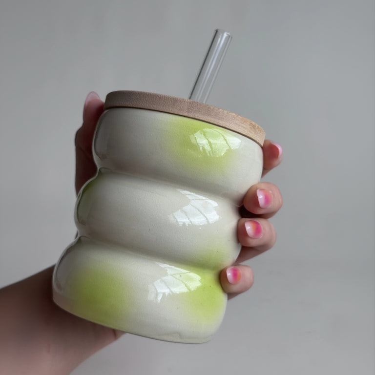 Hand holding a white ceramic cup with a wooden lid and straw on a plain background