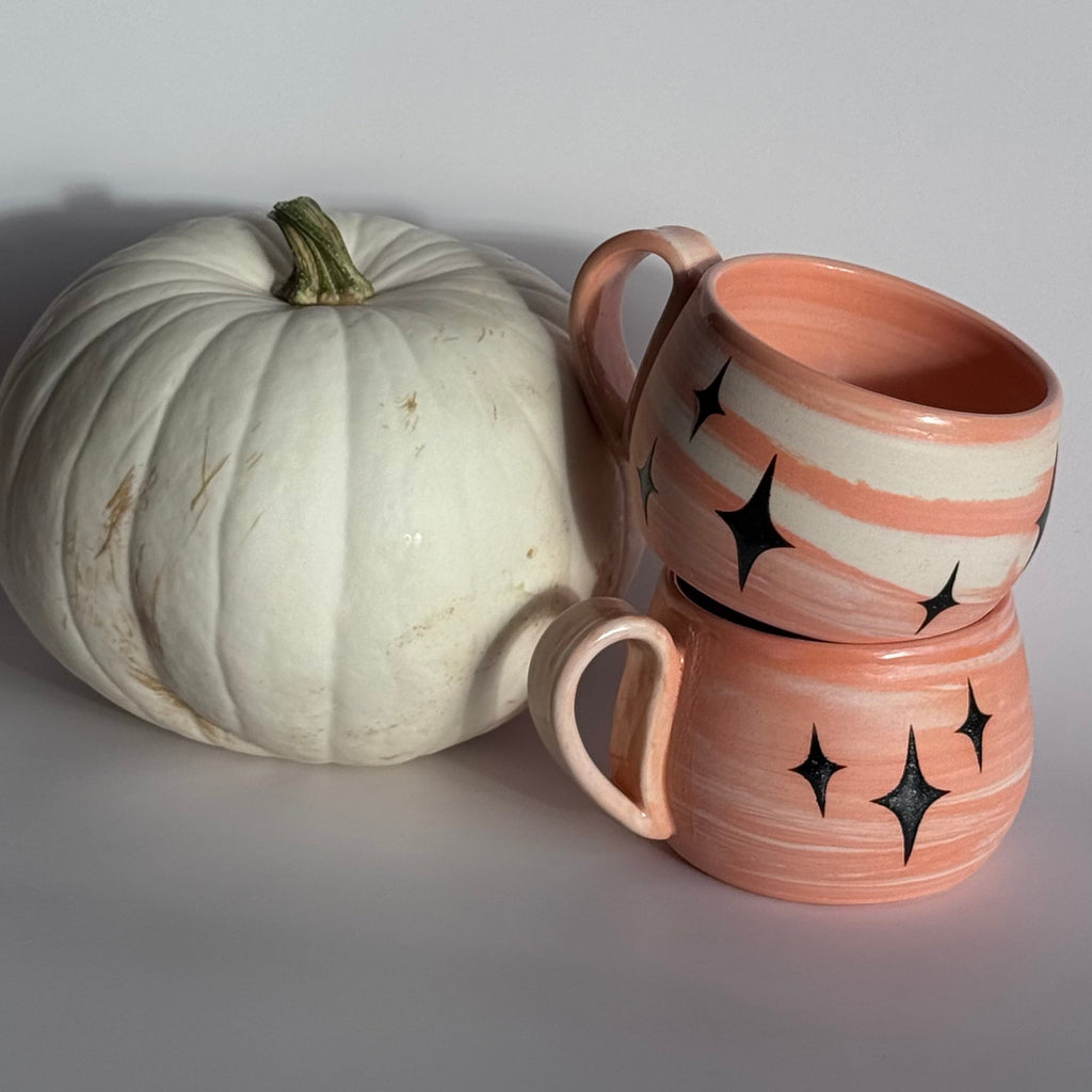 Two pink ceramic mugs with star patterns stacked next to a white pumpkin on a light gray background