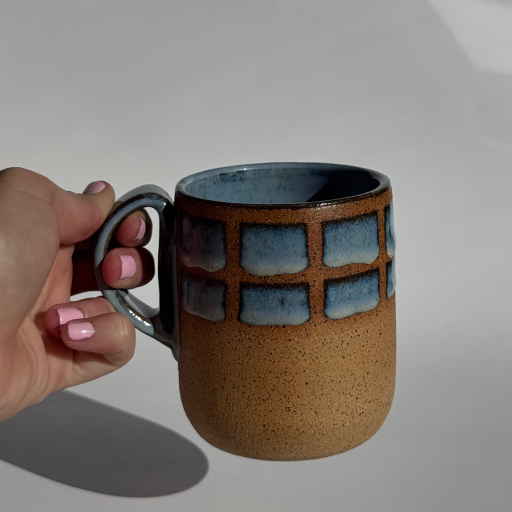 Hand holding a ceramic mug with a textured surface and blue geometric pattern on a light gray background