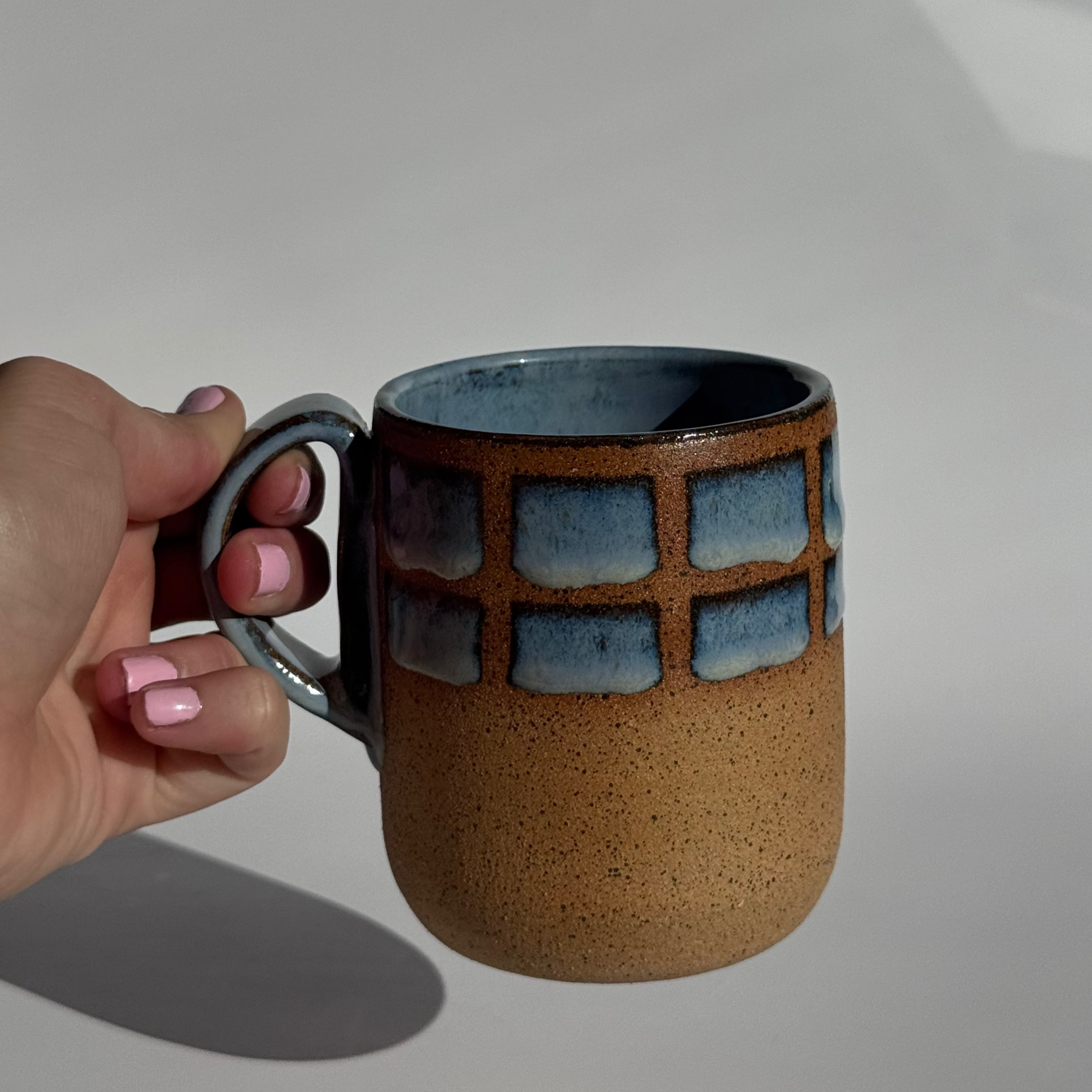 Hand holding a ceramic mug with a textured surface and blue geometric pattern on a light gray background