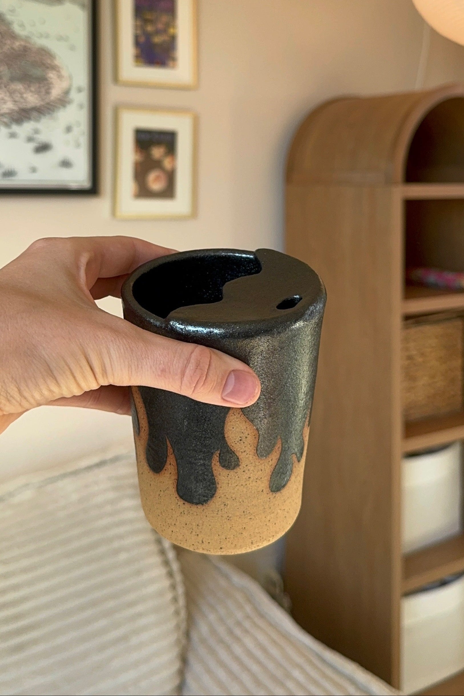 Person holding a ceramic cup with a black interior in a room with a lamp and framed pictures on the wall.
