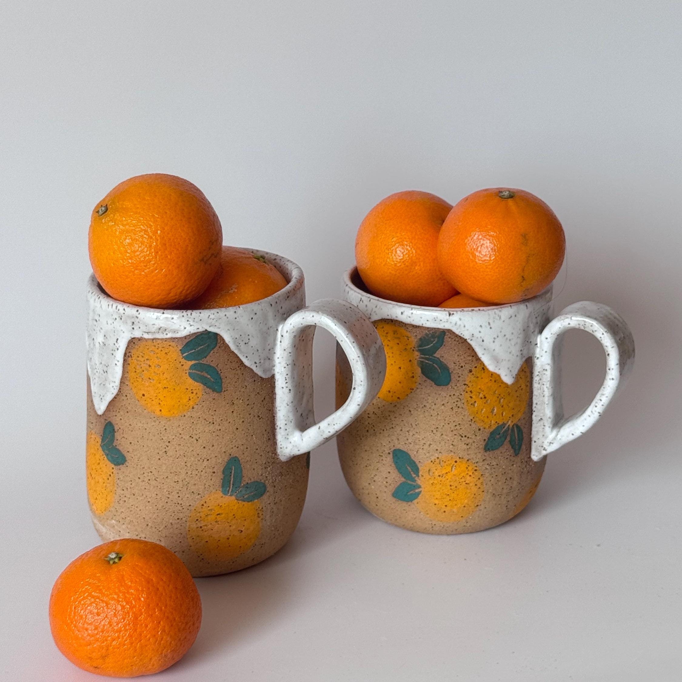 Two ceramic mugs with orange designs filled with oranges on a light gray background